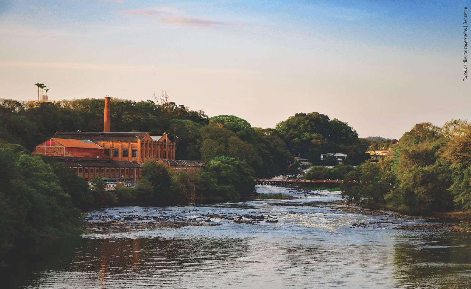 Foto colorida do Rio de Piracicaba e do Engenho Central. O rio largo e de águas escuras ocupa a faixa central da imagem. Á esquerda, os armazéns, galpões, barracões e uma alta chaminé do Engenho Central rodeado de árvores e arbustos. As construções são todas de tijolos à vista. À direita da foto, a mata ciliar à beira rio. Ao longe, a Passarela Pênsil que liga uma margem à outra, o Salto do Piracicaba e parte do Parque do Mirante.
