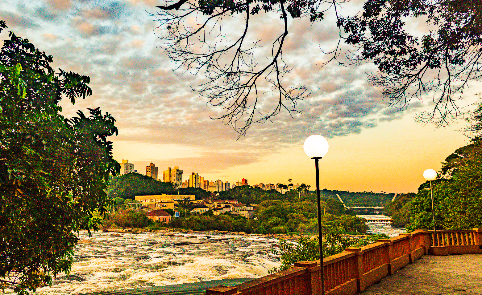 Fotografia colorida da vista de um dos mirantes que compõe o parque. É fim de tarde. Na parte inferior direita, está parte do piso e o guarda-corpos vermelho do mirante. O rio Piracicaba, com águas agitadas, está na parte inferior esquerda, avançando em direção à direita. Ao fundo, a vegetação nativa e alguns prédios. O céu crepuscular que ocupa a metade superior da foto está repleto de nuvens.