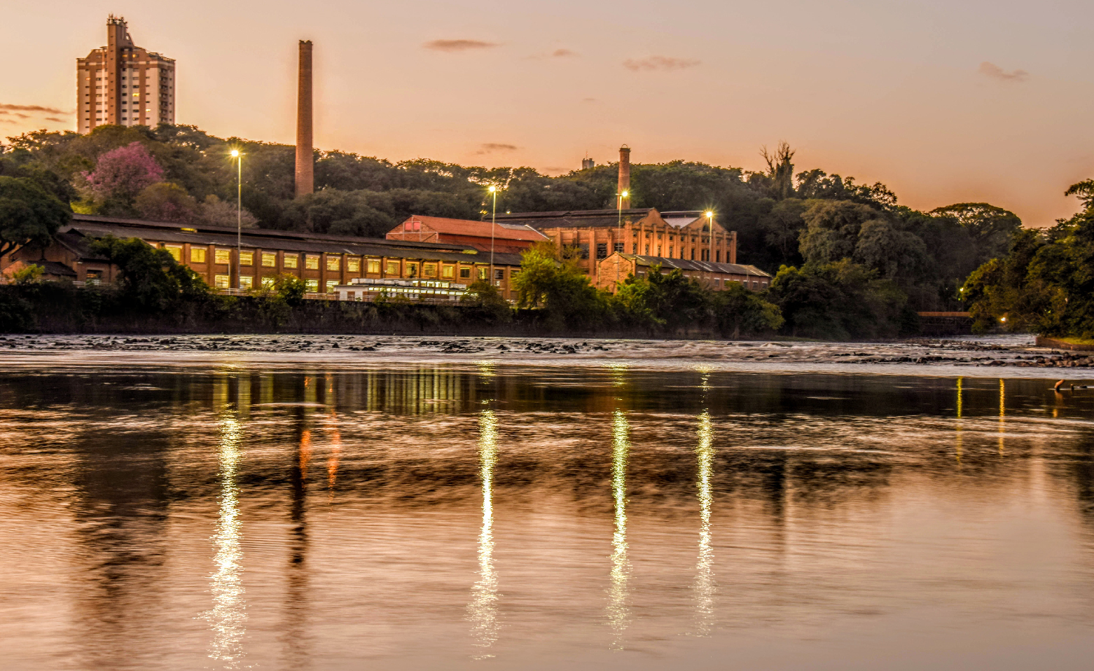 Fotografia colorida do Engenho Central ao entardecer. A foto foi tirada da margem oposta. As águas calmas do Rio Piracicaba ocupam a imagem da metade para baixo. Luzes brilham em cima de quatro altos postes de iluminação pública nos pátios do Engenho Central. É possível ver parte dos altos Barracões Gêmeos, do Teatro Erotides de Campos e do o comprido Edifício 9, além de duas chaminés muito altas. Árvores e arbustos rodeiam todo o Engenho. O céu crepuscular possui tons levemente alaranjados, roxos e cinzas.
