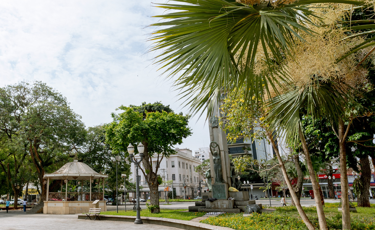 Fotografia colorida da arborizada Praça José Bonifácio. Sobre um canteiro elevado cerca de meio metro do piso cinza, flores, árvores e o Monumento ao Soldado Constitucionalista, com estátuas muito expressivas sobre blocos de cimento. Perto da lateral esquerda da imagem está o Coreto Maestro Osvaldo Peterman.