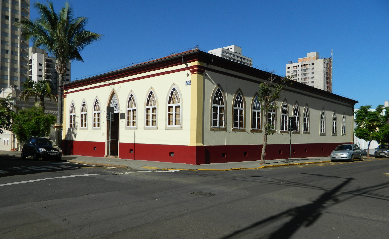 Fotografia colorida das fachadas principal e lateral do Museu, localizado em uma esquina. As paredes são em amarelo claro, com uma faixa horizontal roxa na parte inferior. São repletas de janelas em arcos ogivais. Na fachada principal, há uma porta alta no mesmo estilo. Ao fundo, alguns prédios. O céu está azul e sem nuvens. 