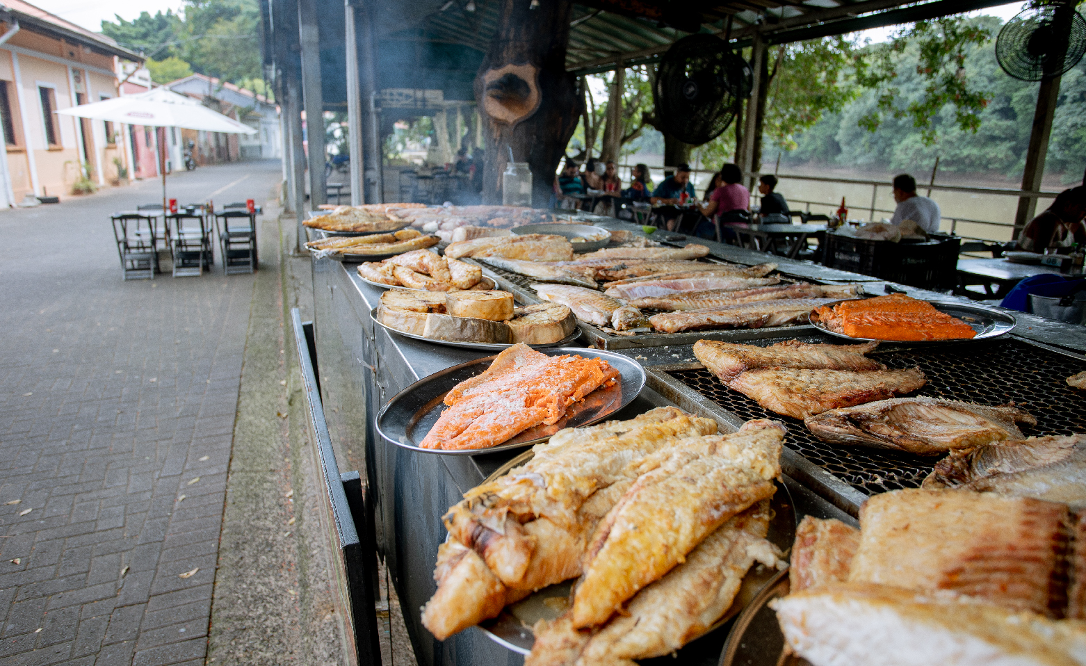 A fotografia colorida mostra peixes sendo assados em um dos restaurantes à beira do Rio Piracicaba. Filés e postas de peixe estão sobre uma comprida churrasqueira que estende-se por todo o lado direito da imagem, até o centro. Ao fundo, em uma área coberta, pessoas comem junto às mesas. No lado esquerdo, está o calçadão de bloquetes da Rua do Porto. 