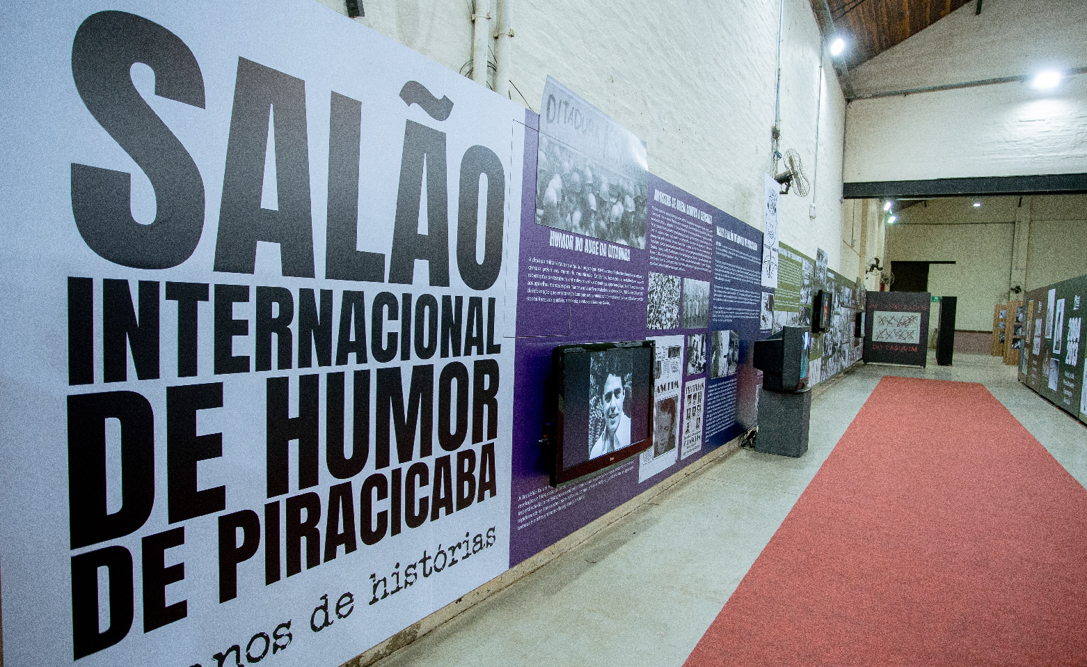 Fotografia colorida. Do lado esquerdo avançando diagonalmente até o centro da imagem, em uma das paredes internas do Barracão 14, há um painel alto e comprido. No lado esquerdo, a inscrição em letras pretas sobre o fundo branco: Salão Internacional de Humor de Piracicaba. Logo ao lado, sobre fundo roxo, textos em letras brancas, fotografias e monitores de TVs. Acompanhando a parede, há um tapete vermelho estendido no piso claro.