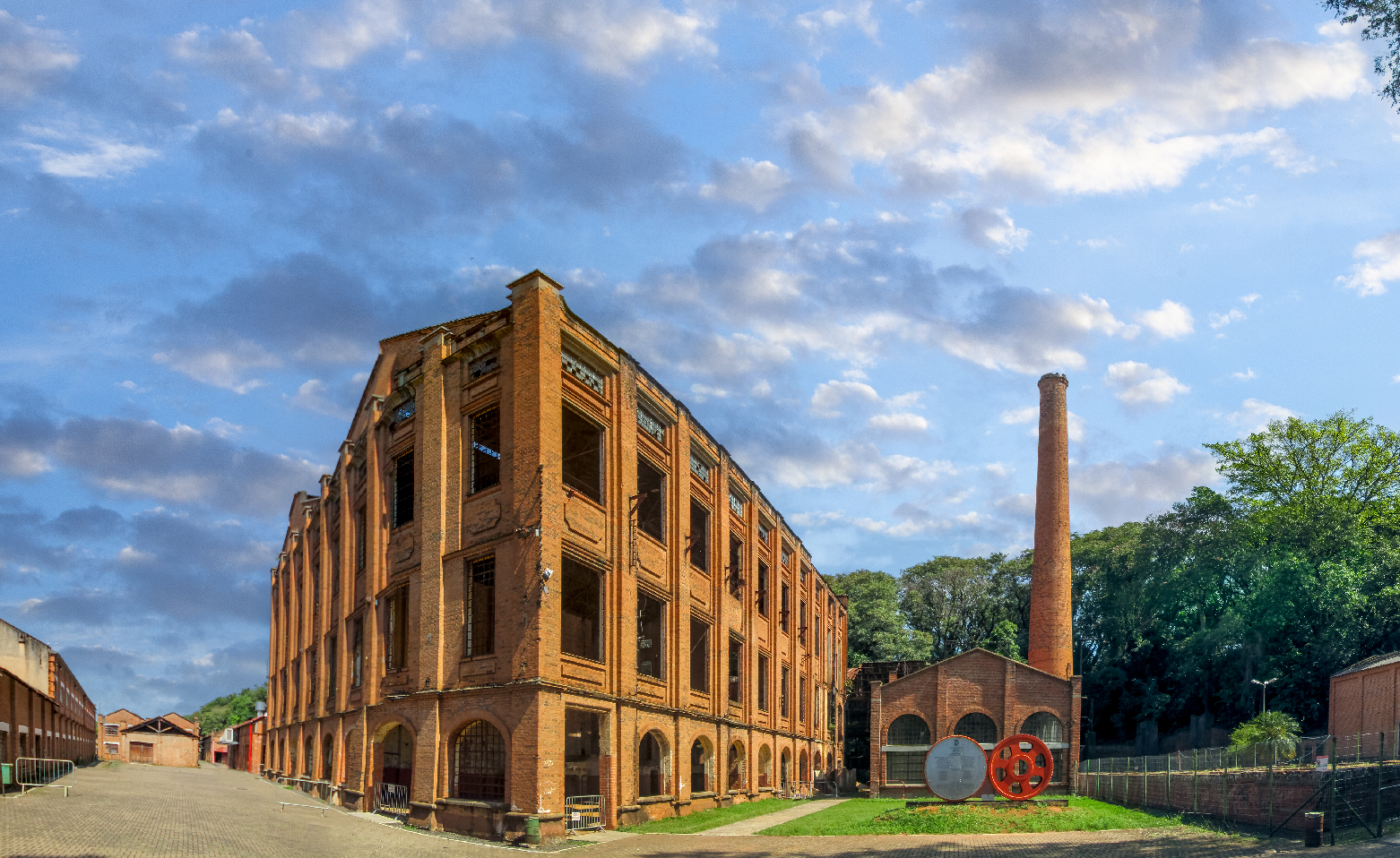 Fotografia colorida. Na parte central, estão os grandes e altos Barracões Gêmeos, construções idênticas e geminadas. São vistas as fachadas e a lateral direita de um deles. São feitos em tijolos à vista e possuem grandes janelas retangulares em três andares. As janelas do térreo são em arco. Do lado direito, há um barracão bem menor, com uma chaminé mais alta que os barracões. O céu azul está repleto de nuvens brancas e cinzas.