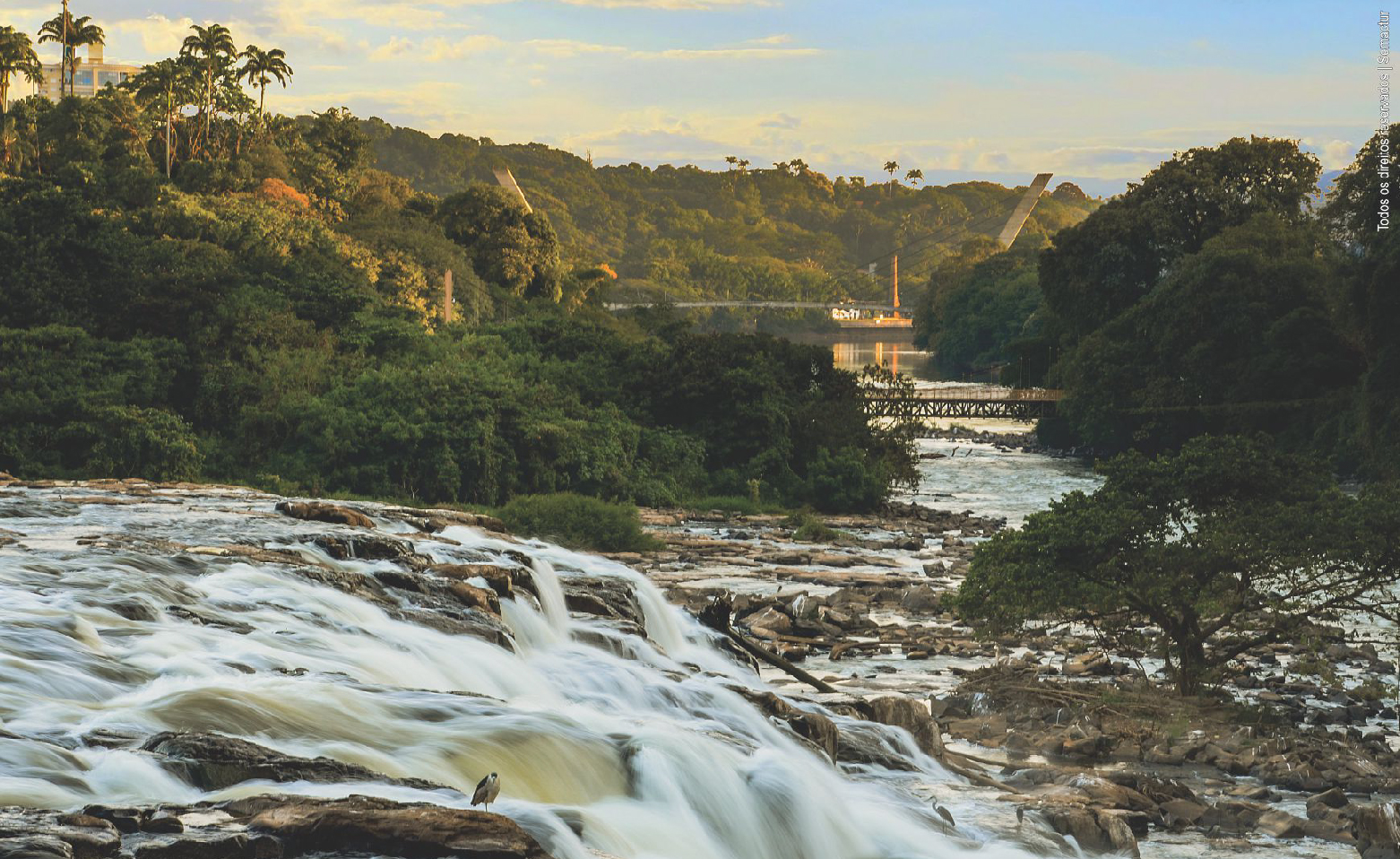Foto colorida do Véu da Noiva tirada na margem direita do Rio Piracicaba, que não está muito cheio. As pedras da corredeira estão aparentes. O movimento rápido das águas está registrado em um rastro ligeiramente borrado. Na margem oposta, arbustos e árvores da mata ciliar. Ao longe, a Passarela Pênsil que liga uma margem à outra.