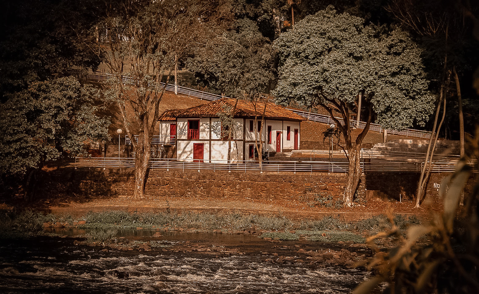 Fotografia colorida da Casa do Povoador, à beira do Rio Piracicaba. A edificação é vista ao longe, na região central da imagem. Tem paredes brancas, portas e janelas vermelhas e está em um patamar superior ao da margem. Há um muro de pedras marrons entre o patamar da casa e a margem. O rio passa na parte inferior da fotografia. Toda a região é arborizada. 