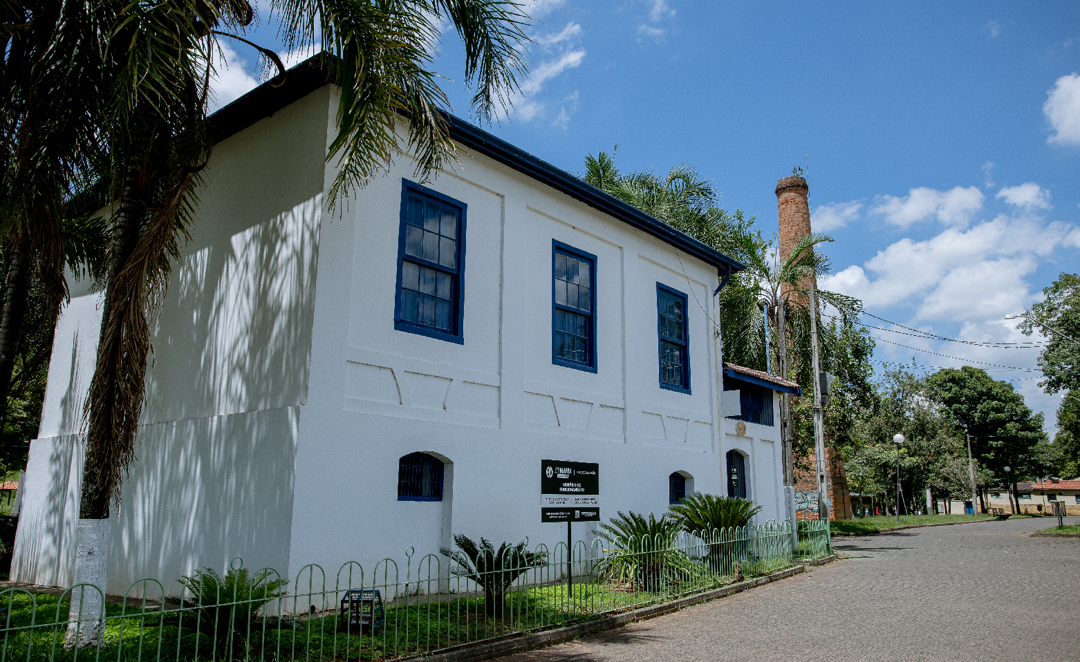 Fotografia colorida da fachada da parte de trás do Casarão do Turismo. A edificação ocupa quase toda a imagem. É um grande sobrado de paredes brancas e janelas e portas azuis. Do lado direito, atrás do Casarão, há uma alta chaminé circular de tijolos à vista. No lado direito da foto, o calçadão de bloquetes da Rua do Porto. 