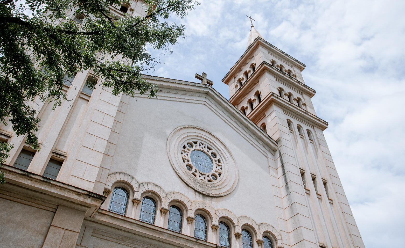 Fotografia colorida registrando a fachada da Catedral de baixo para cima. O céu está repleto de nuvens brancas. As paredes e as duas torres laterais são beges claras. No centro da fachada, há um grande vitral colorido e redondo. Um pouco abaixo, sete janelas em arco lado a lado, também com vitrais coloridos. No topo do telhado de duas águas, uma cruz.