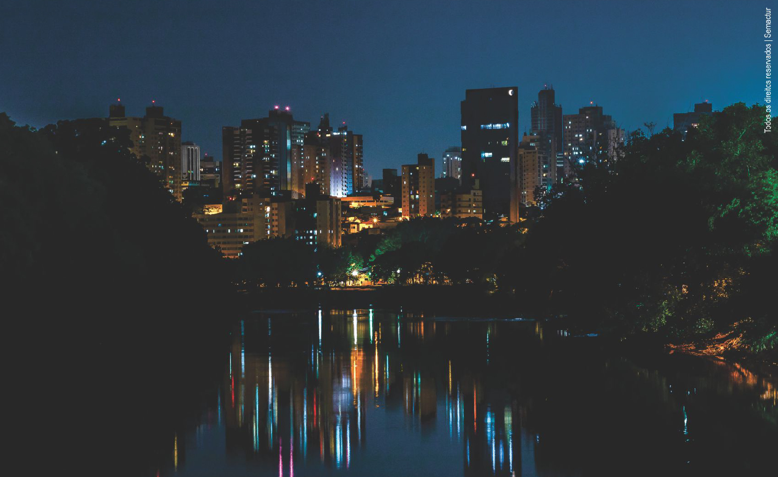 Fotografia noturna e colorida, tirada acima das águas calmas do Rio Piracicaba. Às margens, altas árvores da mata ciliar. Ao fundo vários prédios da cidade com as luzes acesas. O reflexo das luzes e dos edifícios está borrado na água do rio.