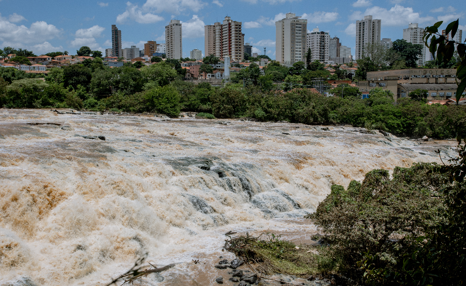  Foto colorida do Véu da Noiva, visto a partir da margem direita do Rio Piracicaba. O movimento das águas escuras correndo nas pedras da corredeira cria uma espuma grossa. Na margem oposta, arbustos e árvores da mata ciliar e, mais ao fundo, prédios e casas da cidade. 