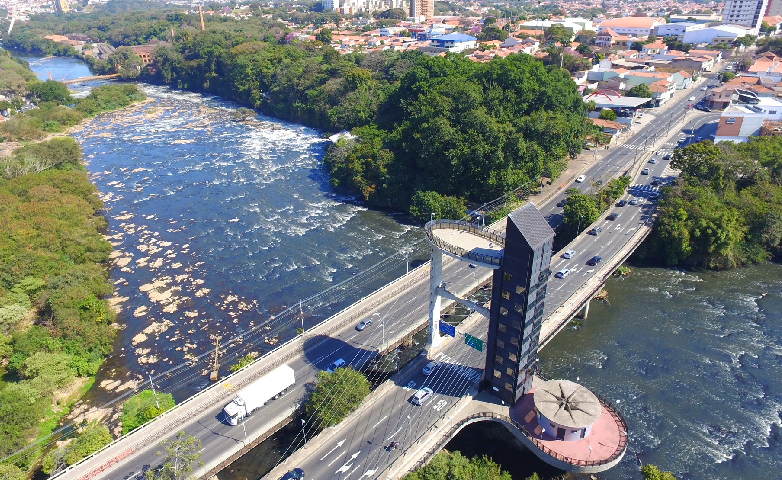 Fotografia aérea e colorida. O Rio Piracicaba ocupa a maior parte da imagem. As águas estão escuras e algumas pedras estão aparentes no leito do rio. Nas duas margens, a densa vegetação nativa. Duas pontes paralelas cruzam a foto do canto superior direito para o canto inferior esquerdo. Na parte central inferior, está a grande estrutura do elevador panorâmico