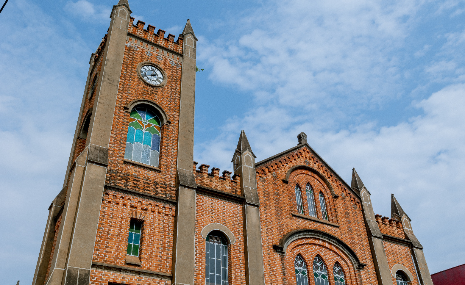 Fotografia colorida da fachada da Igreja Metodista em um dia de algumas nuvens brancas no céu. A foto foi tirada ligeiramente de baixo para cima. A construção de tijolos aparentes possui uma torre alta que está do lado esquerdo da imagem. Perto do topo da torre, há um relógio redondo de ponteiros. Logo abaixo, uma janela em arco com vidros coloridos em tons de azul, verde, marrom e cinza. Há dois tipos de janelas no resto da fachada, a de arcos ogivais, em maioria, e também em arcos. Todas possuem vidros coloridos.