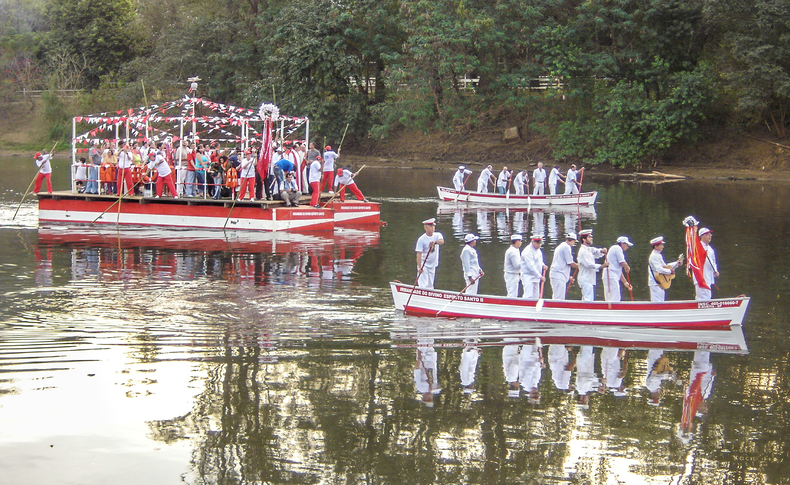 Fotografia colorida que registra uma das cerimônias ligadas à Festa do Divino. Na foto três embarcações lotadas de pessoas navegam pelas águas calmas e esverdeadas do Rio Piracicaba. As duas menores são canoas compridas que estão no lado direito da imagem. Uma mais a frente e a outra ao fundo. Possuem casco branco, com faixas vermelhas pintadas na parte de cima e de baixo. As pessoas em ambas embarcações estão em pé, de roupas brancas e usam compridos remos. Na proa da canoa que está mais à frente, um homem segura um estandarte. Logo atrás, outro homem toca violão. As canoas navegam um pouco à frente da embarcação maior, que está no lado esquerdo da fotografia. É composta por um convés largo, sustentado por duas estruturas compridas que lembram canoas. Pessoas de camiseta branca, calça e boné vermelhos usam extensas varas de bambu para fazer a embarcação avançar. Mergulhando as varas até fincar no leito de lama do rio, elas impulsionam a embarcação. 