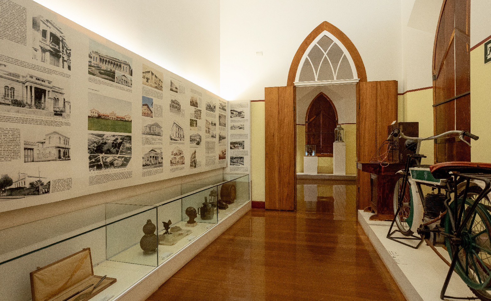 Fotografia colorida de uma das salas internas do Museu. O piso é de madeira. As paredes são amarelas até aproximadamente dois metros e brancas na parte superior. Do lado direito da foto, estão expostas uma antiga central telefônica e uma motocicleta parecida com uma bicicleta. No lado esquerdo, objetos e artefatos variados estão em uma vitrine horizontal. Na parede, há painéis com fotos e textos. Ao fundo, na parte central da imagem, uma grande porta em arco ogival.  