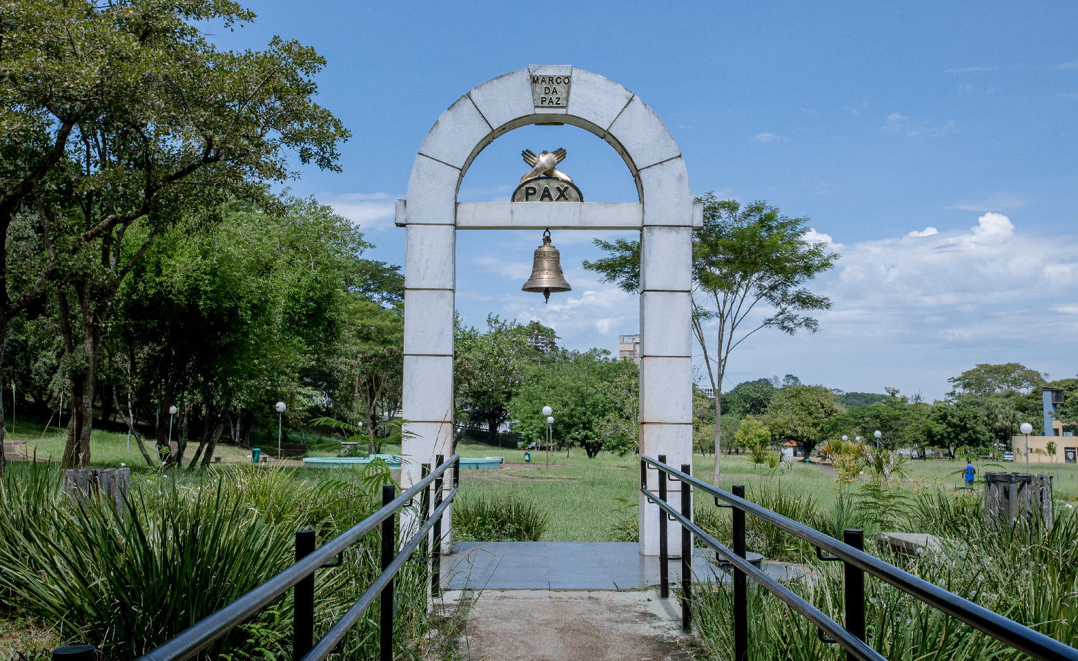 Fotografia colorida do Marco da Paz, visto da rampa com pequena elevação que leva até o monumento. Ao fundo, o grande gramado e, logo após, árvores com copas cheias e verdes. O Marco da Paz é um arco em blocos cinzas. Entre outros detalhes dourados, há um sino pendente. 