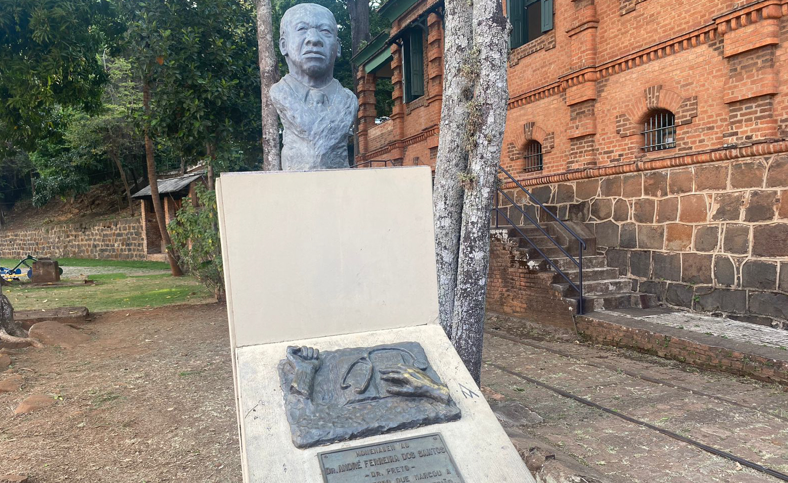 Fotografia colorida do Monumento ao Dr. Preto. A obra foi produzida em pedra de coloração cinza escura e mostra Doutor Preto da barriga para cima, em cima de um totem de pedra. Doutor Preto é calvo, tem olhos amendoados, nariz largo e boca grande com lábios grossos. Um patamar abaixo do busto, em uma chapa de pedra, estão esculpidas duas mãos e uma parte dos antebraços. Entre elas, um estetoscópio está sobre um livro. Ao fundo, uma casa de dois andares de tijolos à vista, um antigo trilho de bonde no pátio, árvores, e um muro de pedras.