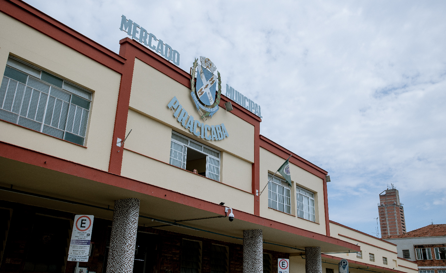 Fotografia colorida do Mercado Municipal de Piracicaba. A fachada é pintada em amarelo claro, com alguns frisos em vermelho. Na parte central da fachada, há um segundo andar, onde estão o letreiro “Mercado Municipal Piracicaba” e o brasão da cidade. Esse andar superior é sustentado por colunas, formando uma marquise em frente à porta da entrada principal.
