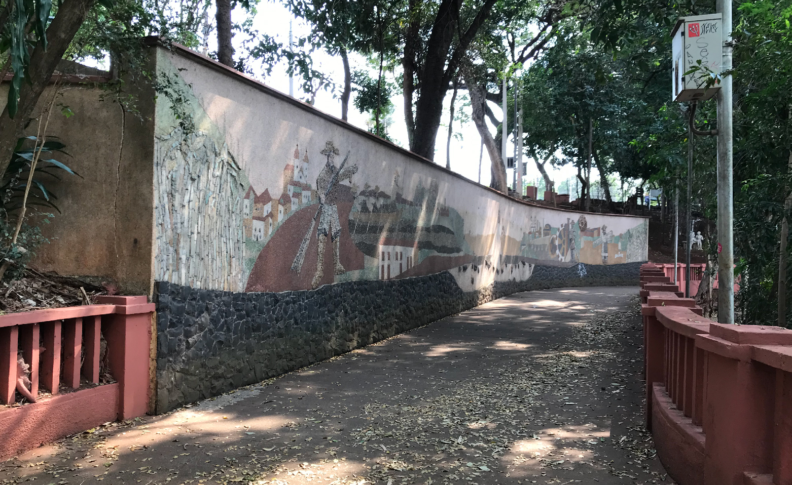 Fotografia colorida que registra, da esquerda para a direita, o grande mosaico de pedras coloridas no muro côncavo, a via para pedestres e a mureta vermelha.