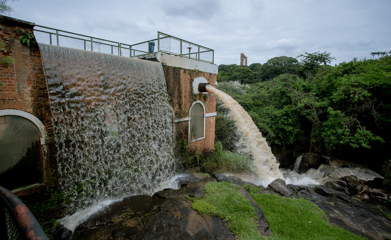 Fotografia colorida da área externa do Museu da Água. Na fachada virada para o Rio Piracicaba, uma cascata cai junto à parede de tijolos à vista, no lado esquerdo da imagem. Um pouco à direita, um grande volume de água jorra de um duto acima de uma janela envidraçada.O museu é cercado por árvores e arbustos. 
