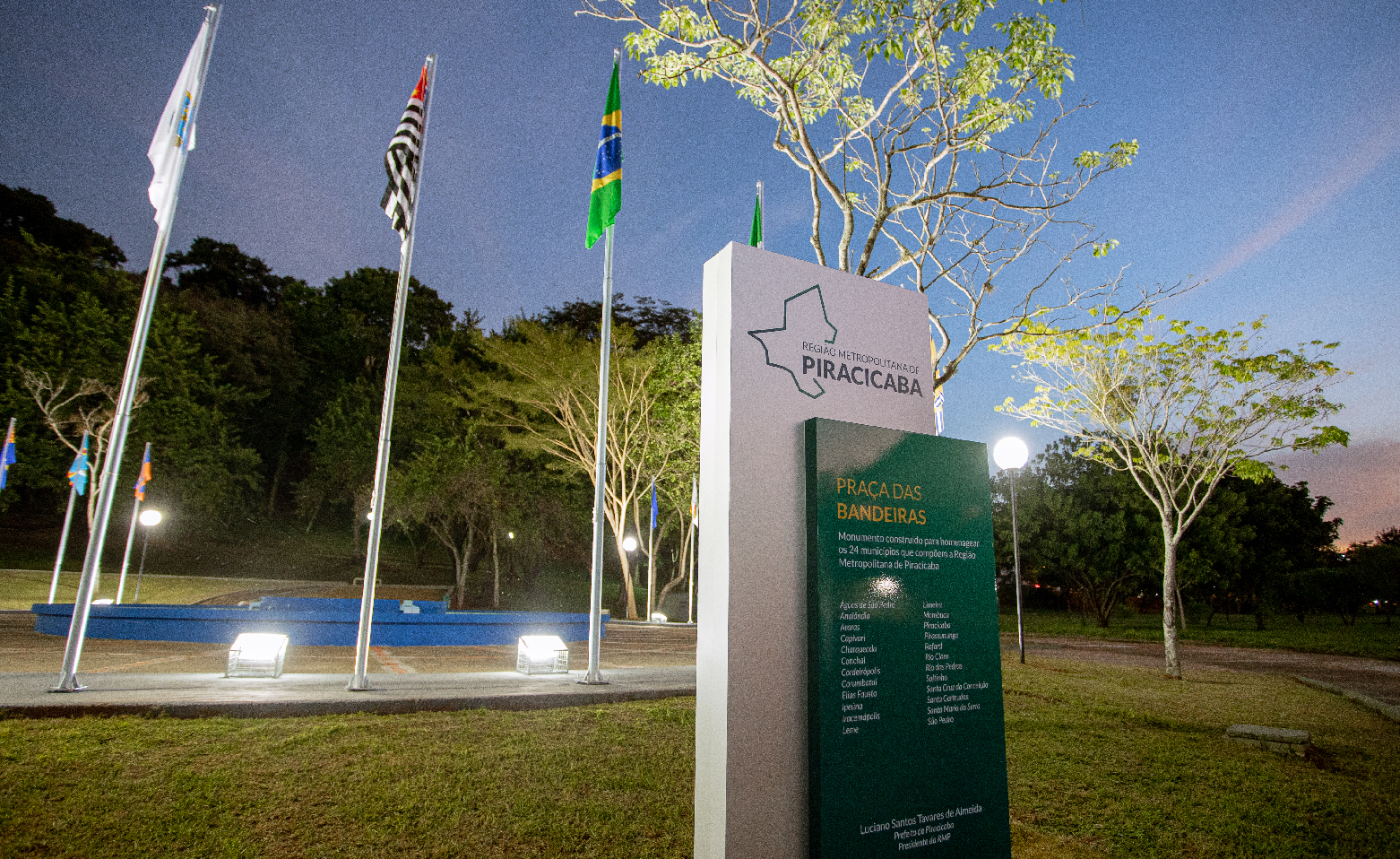 Fotografia colorida ao anoitecer. No centro da imagem, o totem com os nomes das cidades da Região Metropolitana de Piracicaba. Logo atrás, está a praça circular, com um chafariz ao centro e rodeada por um gramado. No chão holofotes virados para cima iluminam as bandeiras no topo de altos e finos mastros. Nesta foto, é possível identificar a bandeira do Brasil e do Estado de São Paulo. O céu crepuscular tem tons de azul até o roxo. 