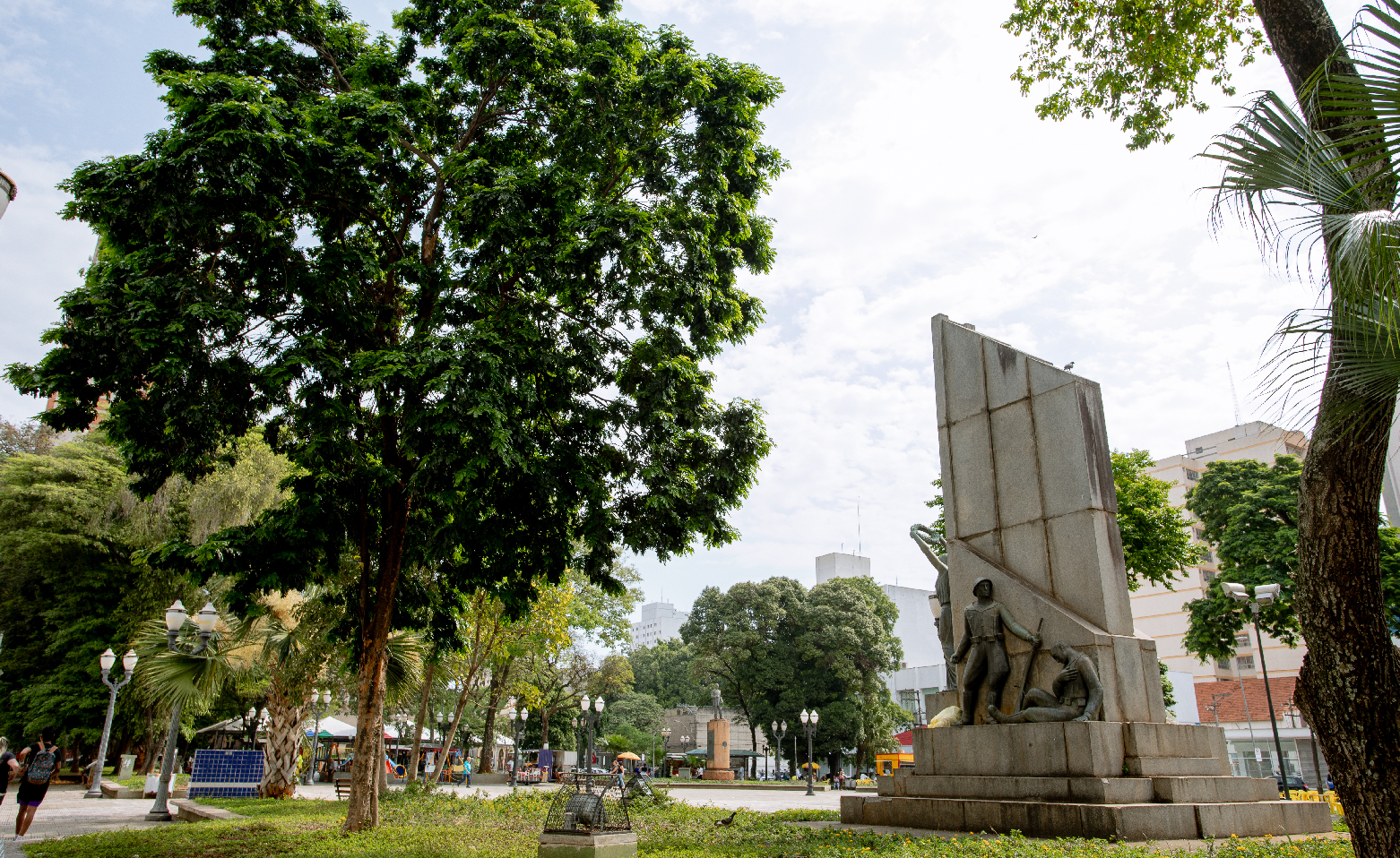 Fotografia colorida de um dos jardins levemente elevados da praça. Sobre o gramado, no lado direito da imagem, está o Monumento ao Soldado Constitucionalista. Do lado esquerdo, há uma árvore alta de copa ovalada e repleta de folhas verdes. Ao fundo o parquinho, postes de iluminação pública, bancos e outros monumentos da praça.