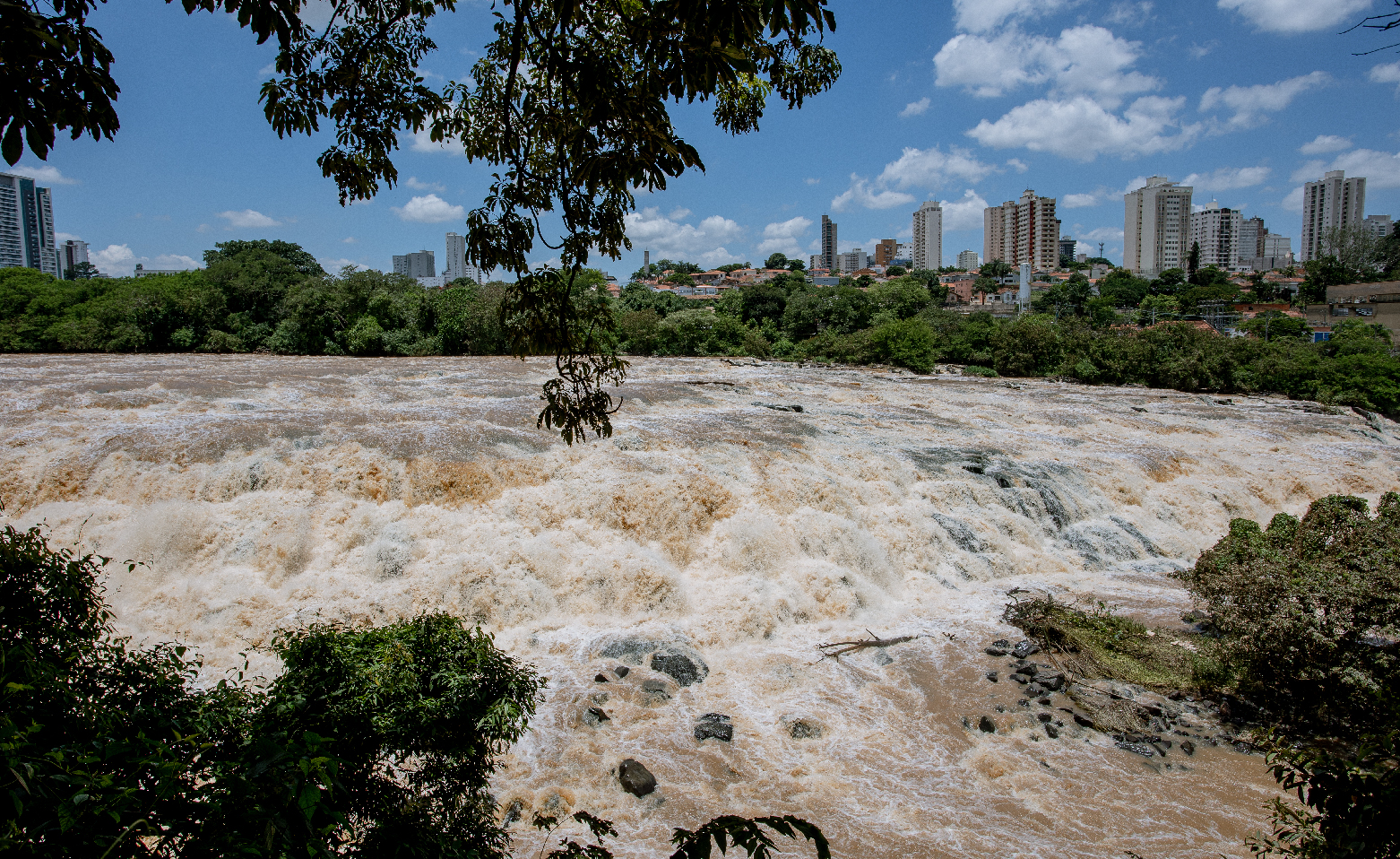 Fotografia colorida da corredeira do Véu da Noiva. Em grande volume, as águas correm encobrindo as pedras e formando a espuma em tons de branco e marrom claro. Ao fundo, a mata ciliar, o topo de alguns prédios e o céu azul com nuvens brancas. 