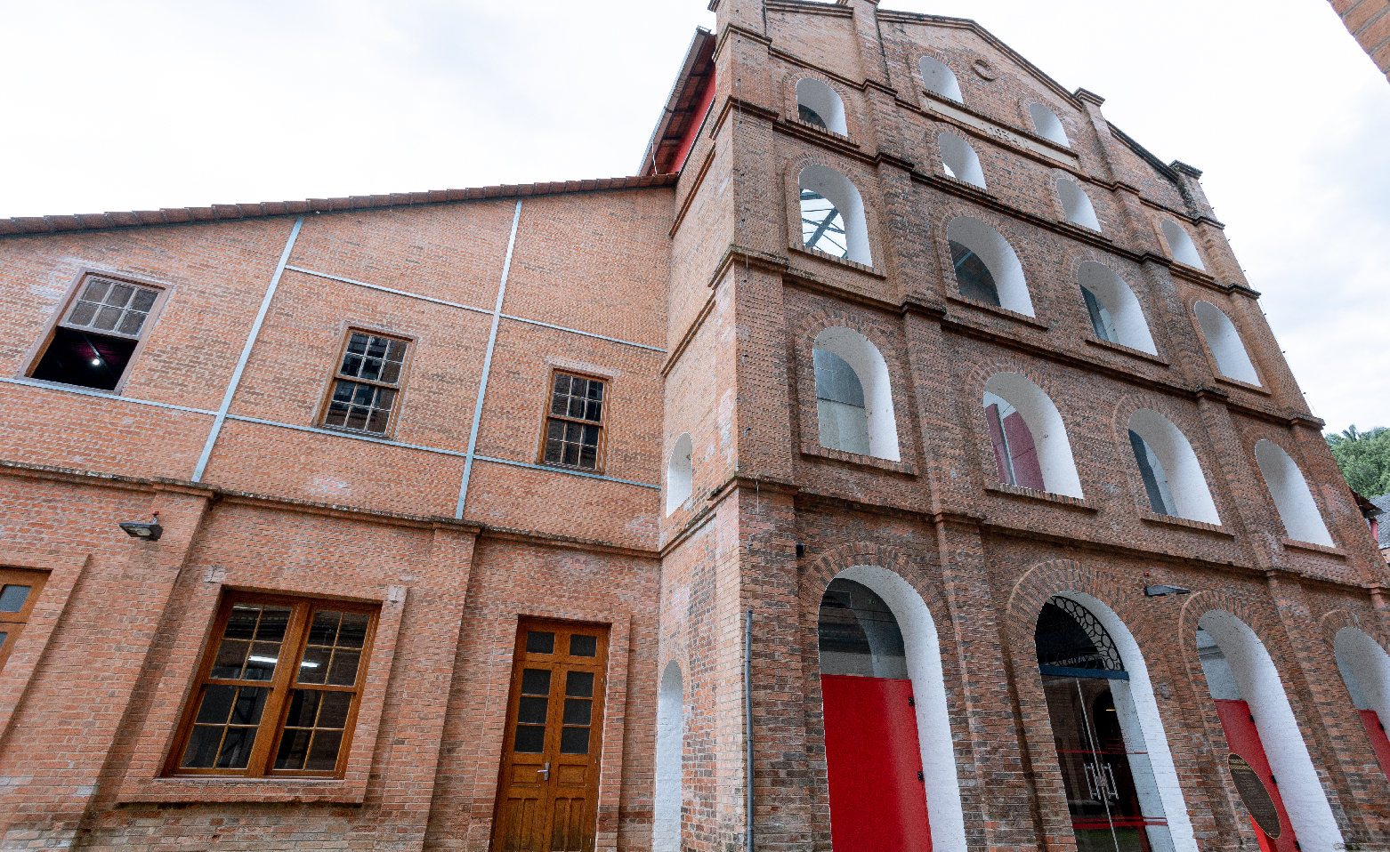 Fotografia colorida e tirada de baixo para cima. Na fachada do Teatro, há quatro passagens em arco lado a lado. A segunda da esquerda para a direita tem a porta de vidro da entrada principal. As outras três são portas vermelhas em metal. Acima de cada arco, existem três grandes janelas, também em arco, uma acima da outra, formando três fileiras horizontais de janelas, lembrando andares de um prédio. Cada janela é fechada por um vidro transparente. No topo, há mais duas janelas em arco. No lado esquerdo da imagem, a porta e as janelas de madeira do café de dois andares. Toda a construção é em tijolos à vista.