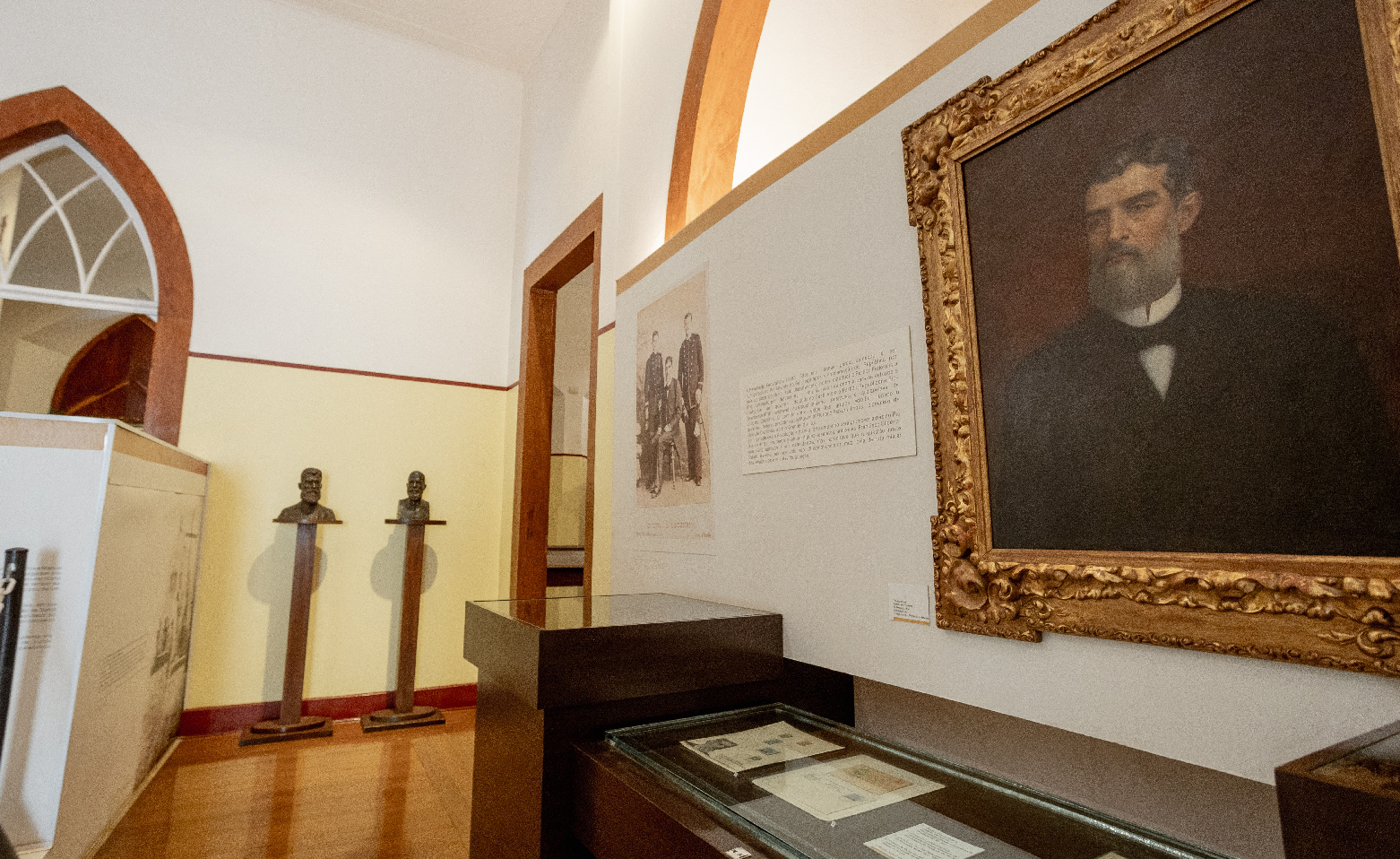 Fotografia colorida. Uma grande pintura com o retrato de Prudente de Moraes está exposta em uma das salas do Museu. O quadro está do lado direito da imagem. Prudente de Moraes é um homem branco, com cabelo curto, barba cerrada e levemente grisalha. Logo abaixo, há documentos expostos em uma vitrine horizontal. Do lado esquerdo da foto, o piso de madeira, paredes e parte de uma porta em arco ogival. 
